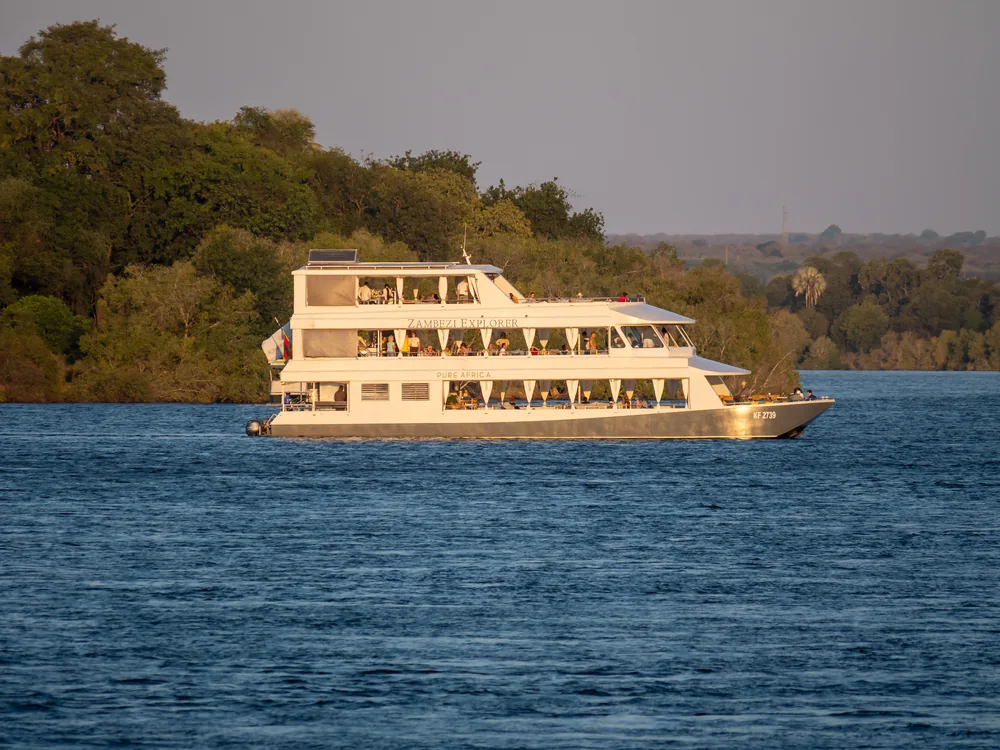 Den tredækkede krydstogtbåd Zambezi Explorer, badet i gyldent lys fra den lave sol, glider forbi skovklædte bredder på Zambezi oven for Victoria Falls