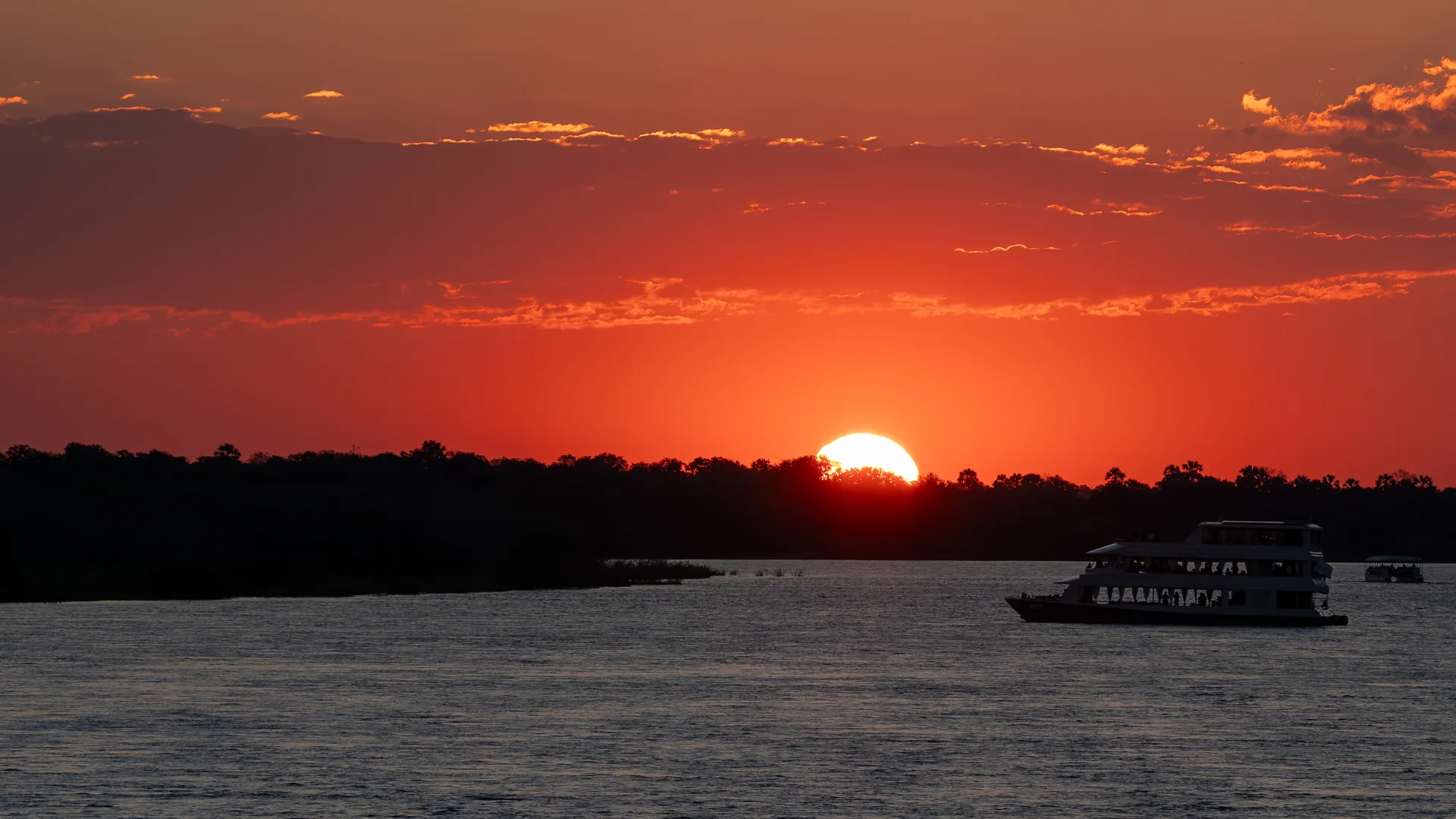 Solen, der går ned i rødt bag trækronerne ved Zambezi-floden oven for Victoria Falls, med en to-dæks cruisebåd i silhuet på vandet