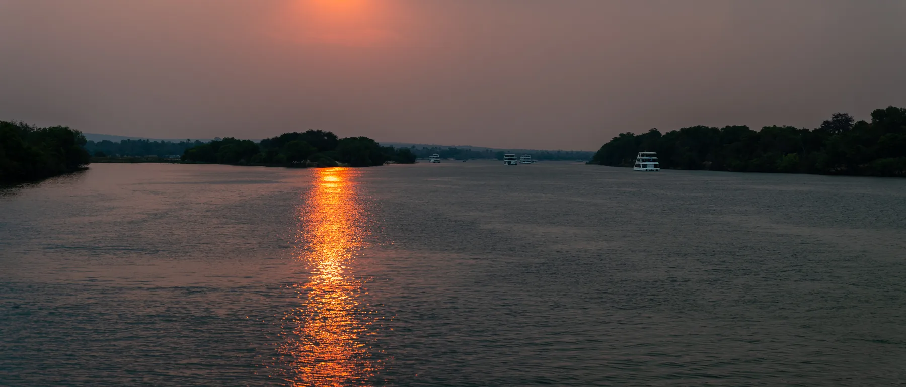 Solen står lavt over en bred, diset strækning af Zambezi i Zimbabwe, og dens spejling løber ned ad vandet mellem skovklædte øer