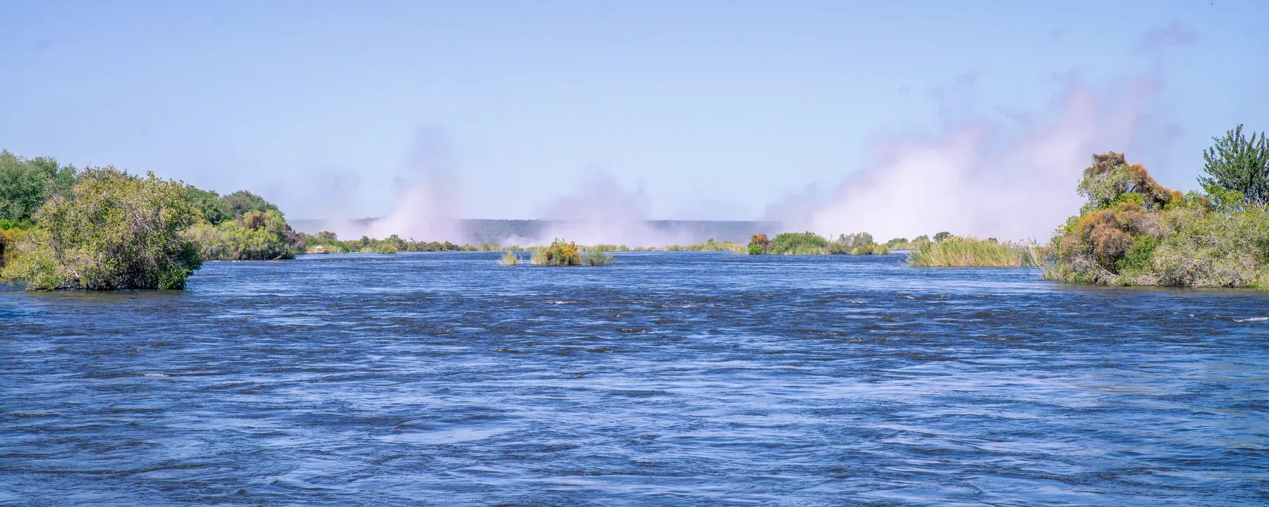 Vandtågen fra Victoria Falls stiger op bag træklædte øer i Zambezi-floden opstrøms for faldet